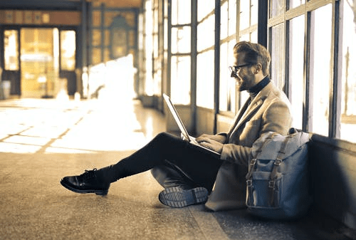 a person using a laptop while sitting on the ground with his carry-on baggage beside him