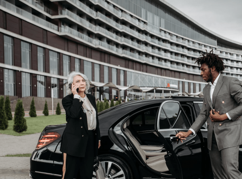 An image of a man in grey suit opening the car door for a woman in a black suit