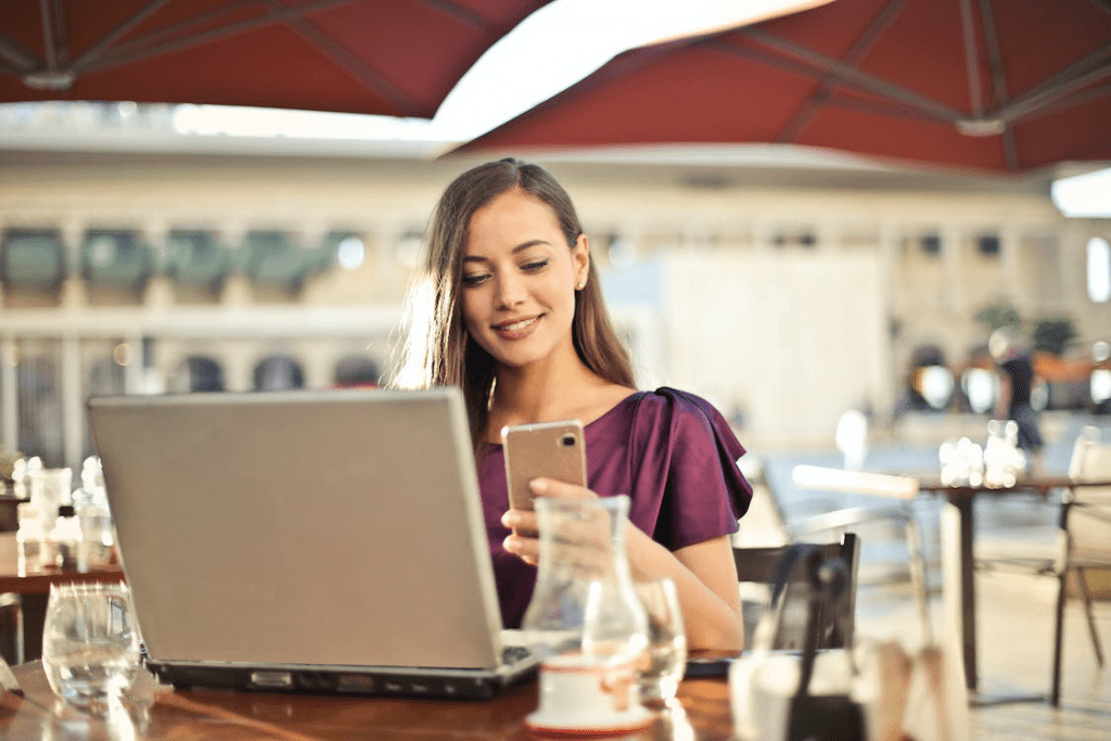 An image of a woman sitting on a chair wearing a purple dress while looking at her phone and working on her laptop