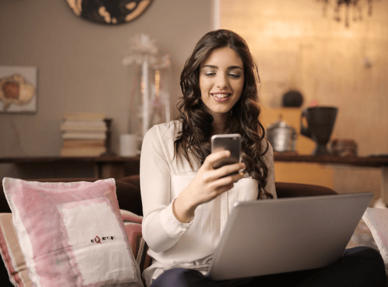 An image of a woman sitting on the sofa while looking at her phone and working on her laptop in her lap