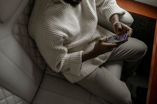 An image of a man wearing a white sweater and holding his phone while sitting in a car